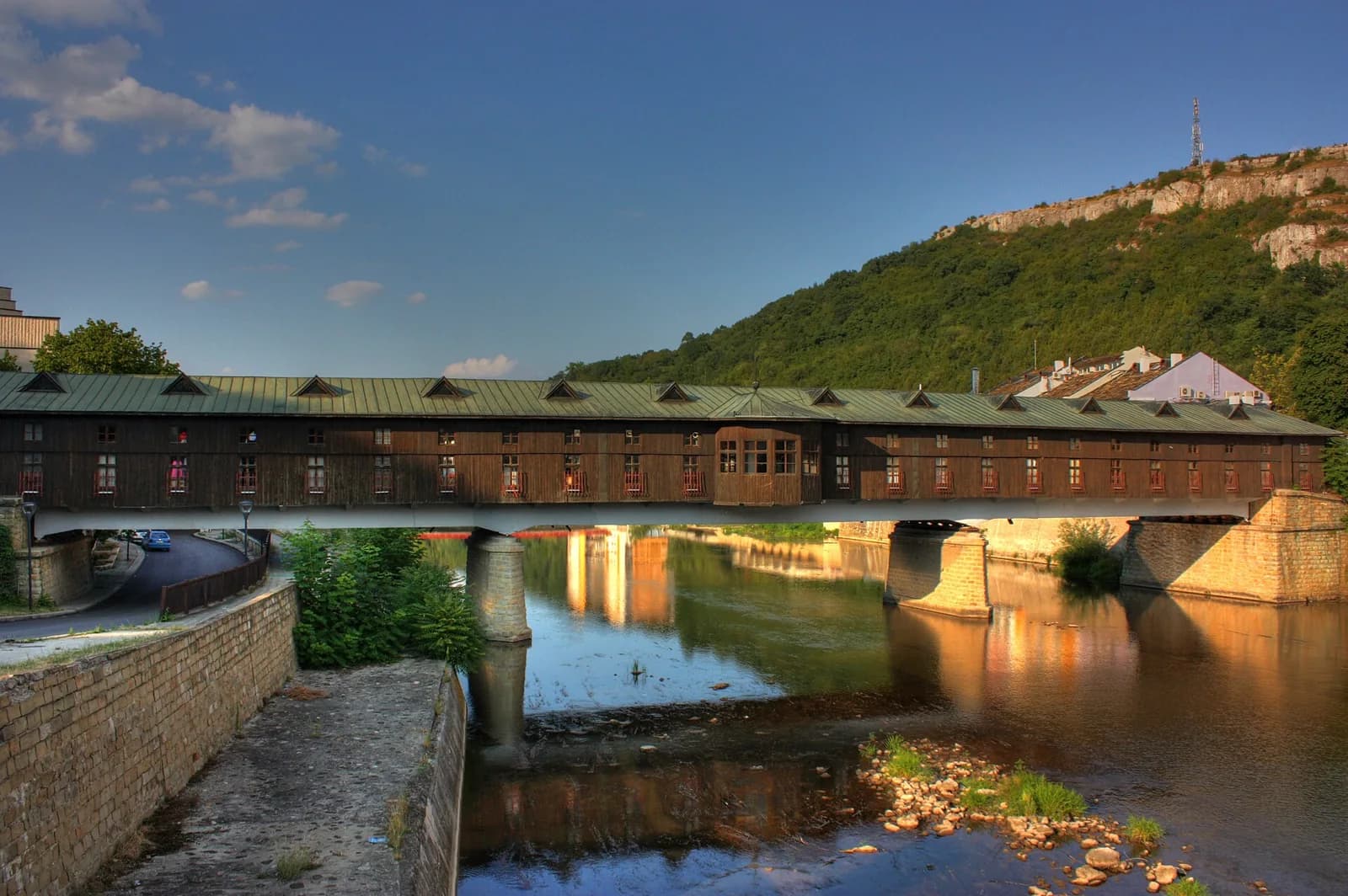 Lovech's timber-clad Covered Bridge with a green metal roof over the Osam, with a wooded limestone hill behind