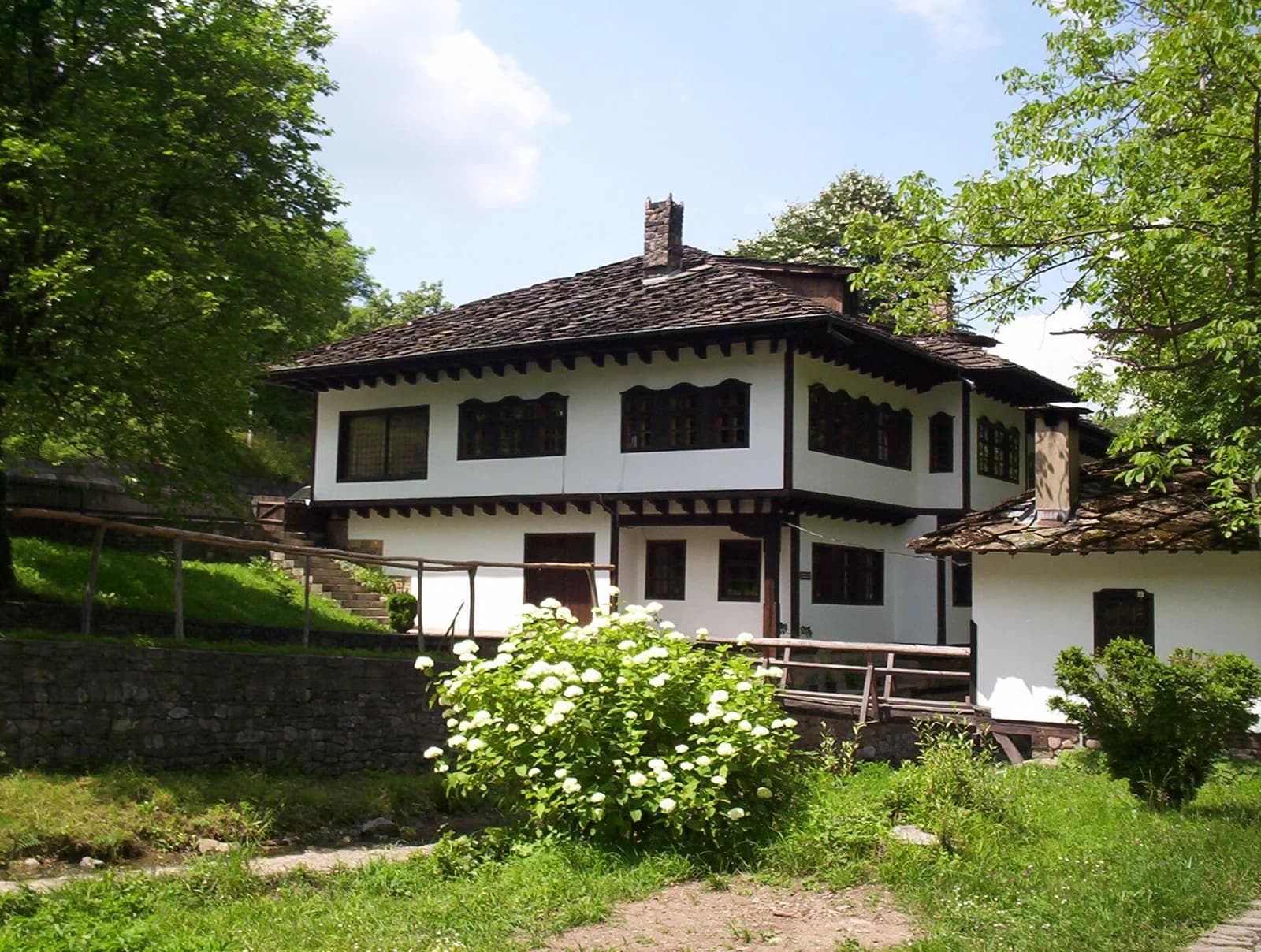 A whitewashed two-storey Revival-period house with dark timber trim and a stone-slab roof among trees at Etar