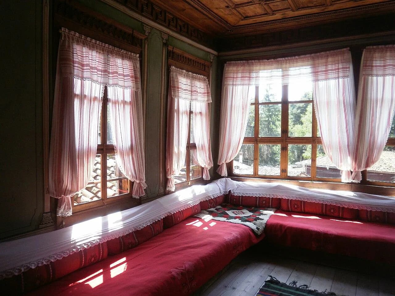 A sunlit corner with curtained windows and a red upholstered bench in Oslekov House.