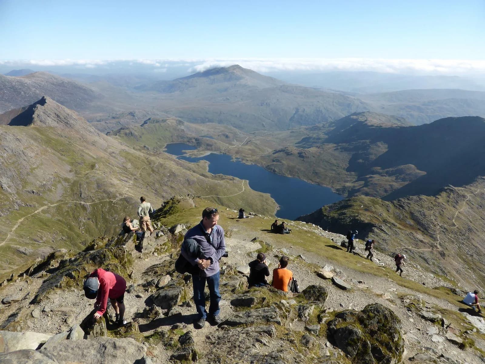 Hikers on a rocky summit path above Llyn Llydaw and distant ridges on Yr Wyddfa.